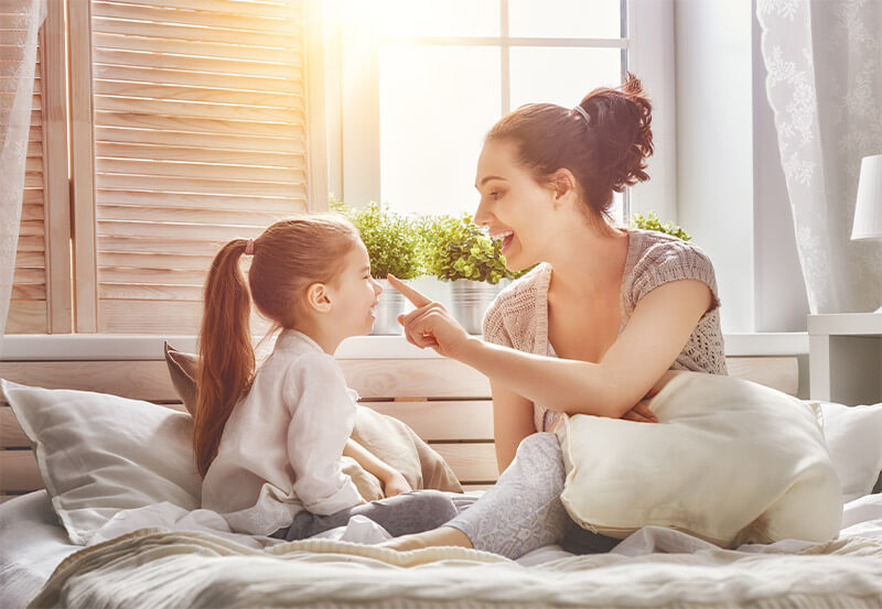 A mother and daughter play on a bed while the sun beams through the bedroom window.